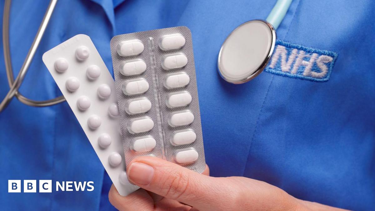 Close up of a hand holding two packets of white tablets. A blue nurses top is in the background with the letters NHS written on in white
