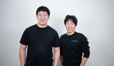 Two men in black t-shirts stand against a gray background.