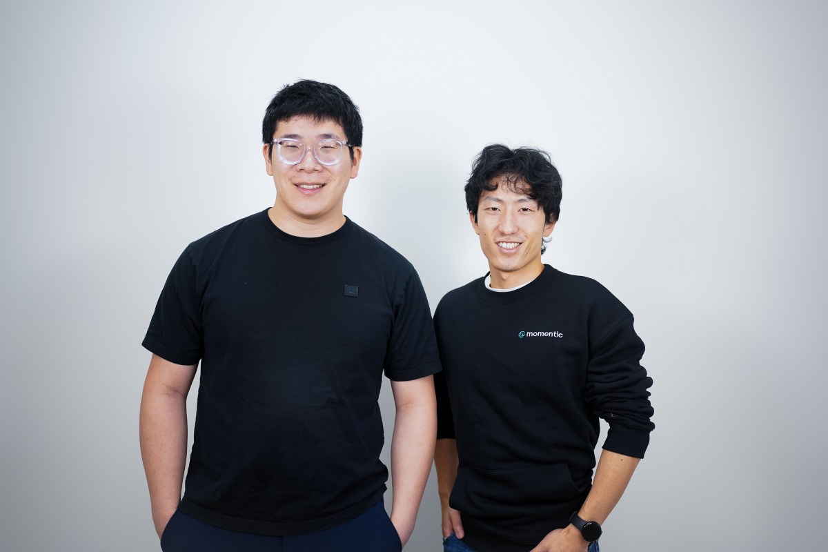 Two men in black t-shirts stand against a gray background.