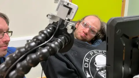 Close-up of assistive technology equipment with flexible arms and a mounted device positioned near a person wearing a black hoodie featuring a circular logo with the text "DR PEACE". A tablet screen is visible in the foreground, and a bright green wall forms part of the background.