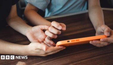 Two children, their faces not visible, hold a smart phone in an orange case on a brown table top.
