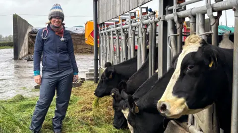 BBC A vet stands in a farmyard.  She is dressed in a navy top and waterproof trousers with a colourful bobble hat. She has a stethoscope around her neck and a wide smile. Five cow heads poke through the bars of their shed to reach some grass on the ground. The dairy cow biggest in frame is side-eying the camera. 