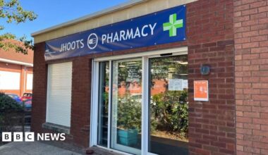 The front of the Jhoots branch in Victoria Square, Portishead in bright sunlight. Above the door a giant blue banner featuring NHS branding reads "Jhoots Pharmacy". To the left of the door a window has shutters pulled down in front of it.