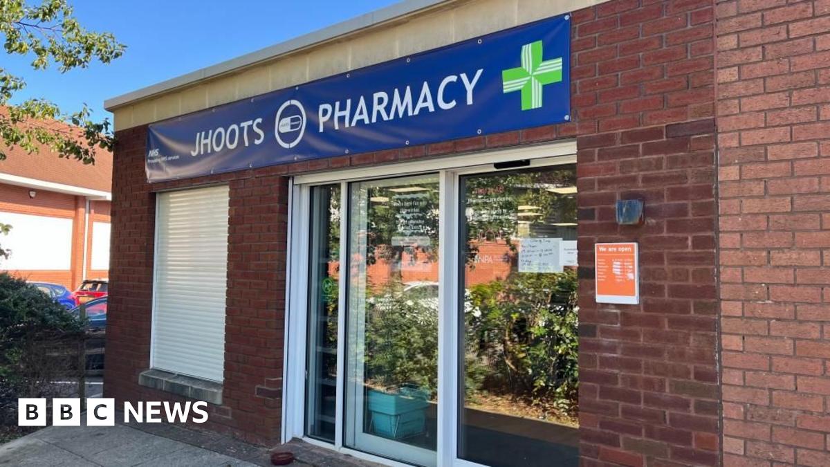 The front of the Jhoots branch in Victoria Square, Portishead in bright sunlight. Above the door a giant blue banner featuring NHS branding reads "Jhoots Pharmacy". To the left of the door a window has shutters pulled down in front of it.