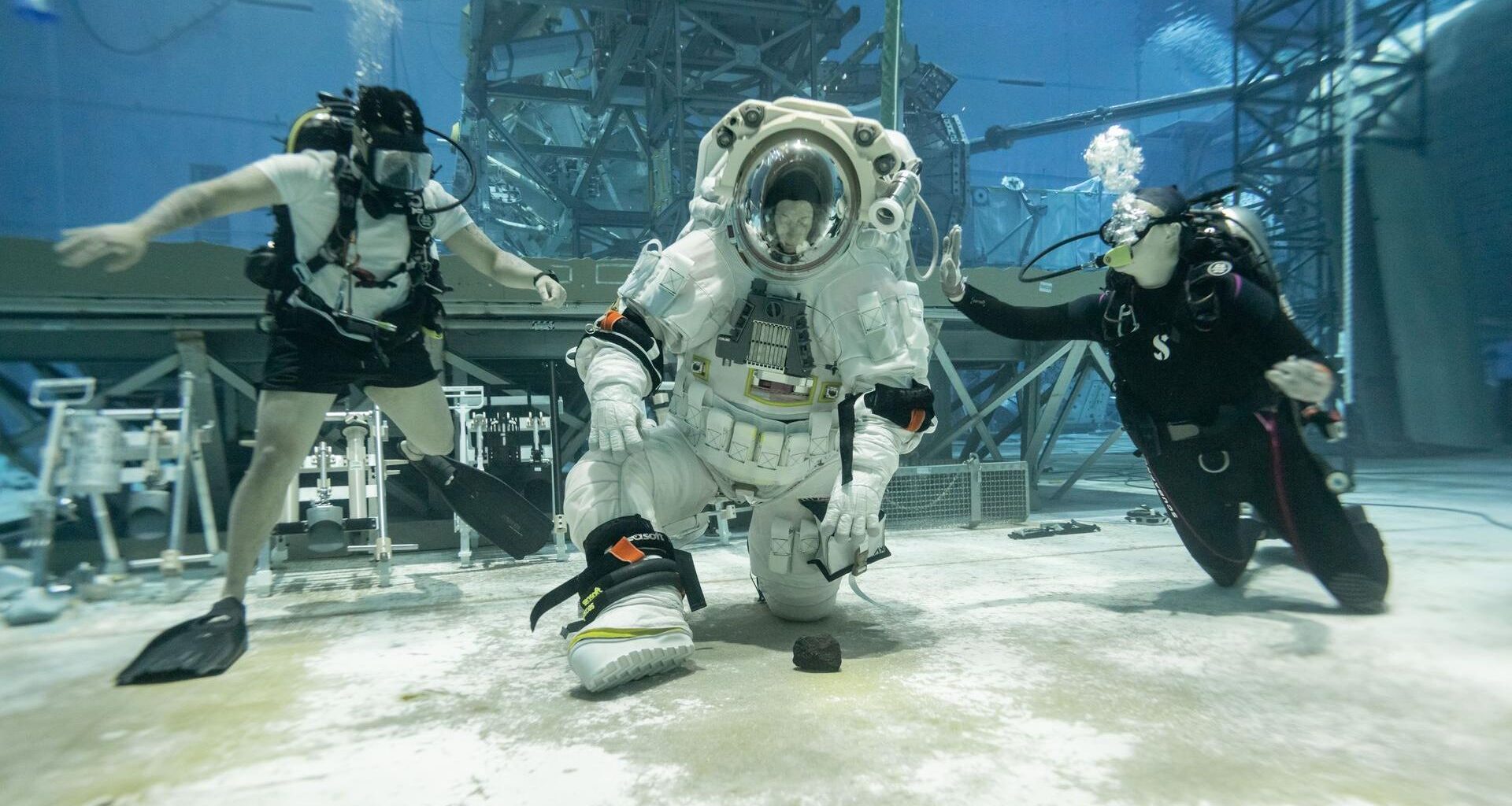 A person wearing a white spacesuit with a clear helmet kneels down to pick up a rock at the bottom of a large sandy pool with two people wearing dark swimsuits on either side of them