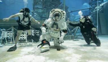 A person wearing a white spacesuit with a clear helmet kneels down to pick up a rock at the bottom of a large sandy pool with two people wearing dark swimsuits on either side of them