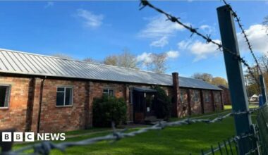 A brick building. It is sunny. Barbed wire is in front of it.