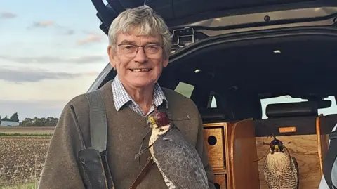 Contributed Mick Smith smiles at the camera as he stands at the back of vehicle with it boot open. A peregrine falcon with a cap over its head rests on his arm. He has grey hair and wears a green jumper with a shirt underneath. 