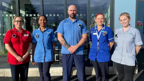 WHSCT A group of five health care staff wearing uniforms stand in front of a hospital building. A man is at the centre of the image, the other four are women.