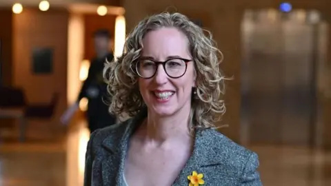 Getty Images Lorna Slater - a woman with curly blonde hair down to her shoulders, wearing glasses and a grey suit jacket with a yellow daffodil on the lapel - pictured smiling at the Scottish Parliament