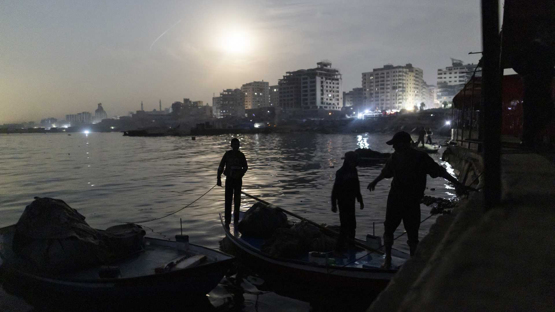A coastal view of Gaza City, showing the sea meeting the shoreline at night. The moon is visible as a bright blur of light shining above the city skyline, while boats are visible moored to a dock in the foreground, being worked by local people.