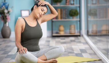 woman sitting crossed legged on a yellow mat on a grey lino floor tilting her head to the side and holding it with one hand, eyes closed