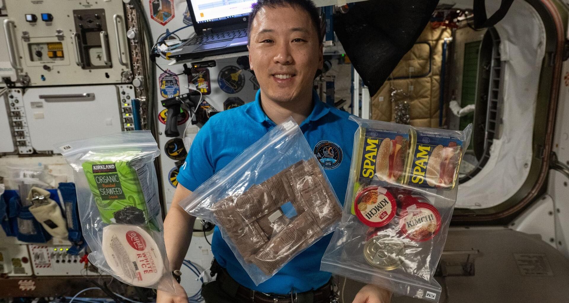 A man with short black hair wearing a dark blue shirt floats in microgravity behind various plastic covered food packages.