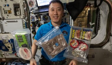A man with short black hair wearing a dark blue shirt floats in microgravity behind various plastic covered food packages.