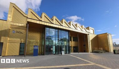 A multi-storey building with a shiny gold exterior and a large glass entrance is photographed on a sunny and largely clear day.