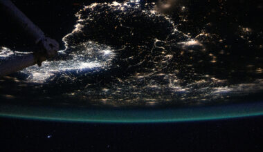 Earth is a dark sphere with a glowing green border in this photo. We can only see one portion of it; in this photo’s orientation, we’re looking at the “bottom” of Earth’s sphere. City lights shine brightly at middle left; they are more dispersed throughout the rest of Earth that’s visible. Part of the space station is just barely visible at top left.