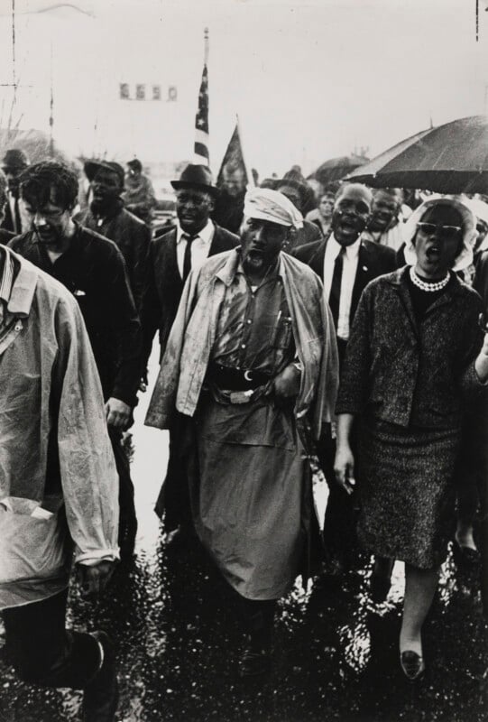 A group of civil rights activists marches down a wet street, some carrying flags and others holding umbrellas. The group includes men and women, wearing formal and casual clothing, walking together with determined expressions.