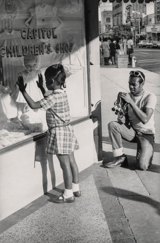 A young girl in a plaid dress looks through a children's shop window, hands pressed to the glass, while a man kneels nearby on the sidewalk, adjusting a camera with a busy street in the background.