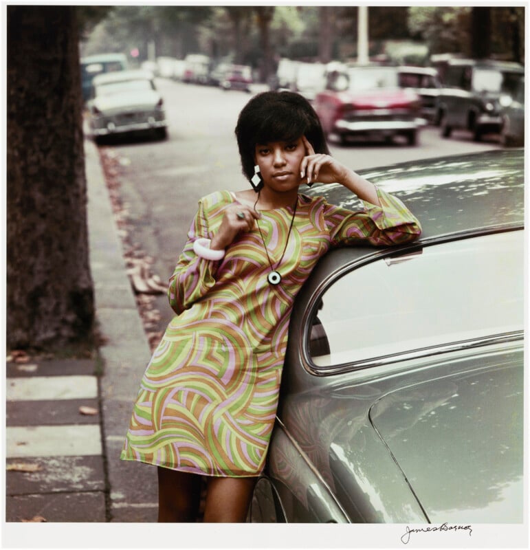A young woman in a colorful, patterned mini dress leans against a vintage car parked on a tree-lined street, with parked cars and blurred traffic in the background.