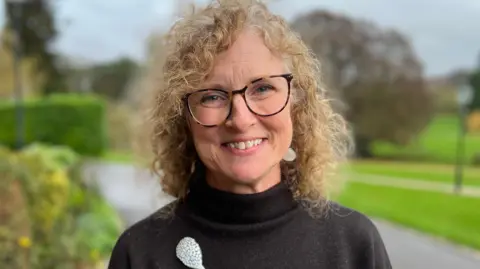 A person wearing a black turtleneck jumper with two white floral designs stands outdoors on a paved path. The background includes greenery, shrubs, and trees with autumn foliage, along with a stone building partially visible on the left.