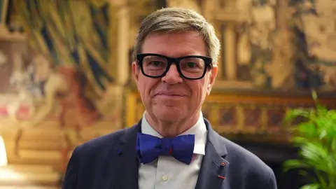 Getty Images A close-up of Yann LeCun wearing thick rimmed black glasses, a navy suit with a dark blue bowtie standing in an opulent room at Buckingham Palace.