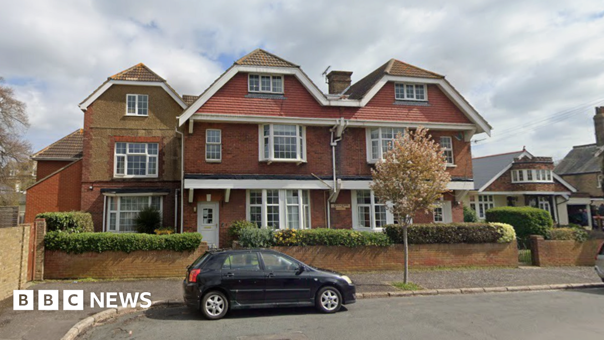 A large three-storey building made of red and yellow bricks, with a roof pointed in three places and large, white windows. A small black car is parked on the road in front of the property.