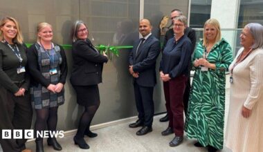 Smartly dressed people stood in a line, smiling at the camera. A woman in the middle is cutting a green ribbon over a grey door using a large pair of scissors.
