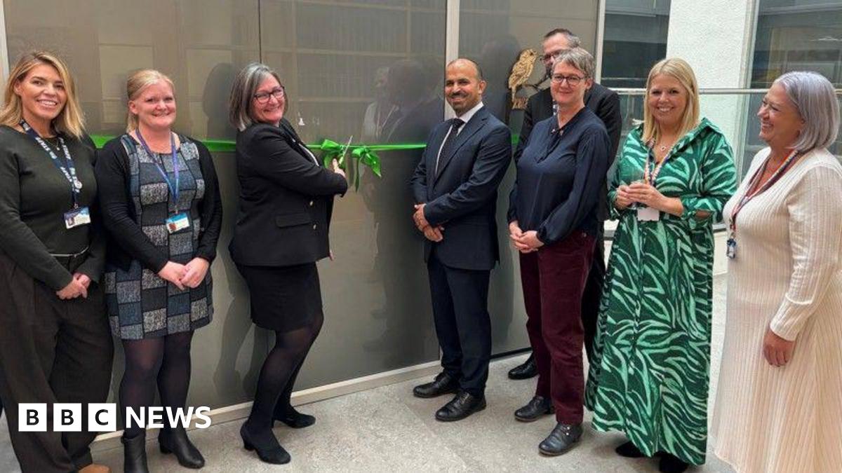 Smartly dressed people stood in a line, smiling at the camera. A woman in the middle is cutting a green ribbon over a grey door using a large pair of scissors.