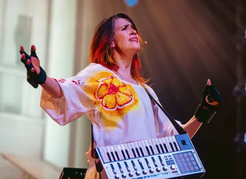 Getty Images Imogen Heap performs on stage in Los Angeles earlier this year. She has her arms outstretched and is wearing a white top with an orange flower on it,, and black gloves.