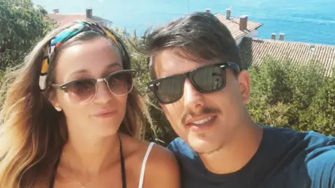 Grenfell Love Foundation Young couple, one woman with medium brown hair and a young man, with short dark brown hair and a moustache both wearing sunglasses and looking at the camera taking a selfie. She has thin straps for a top and he is wearing a blue t-shirt - behind them are rooftops and the sea. 