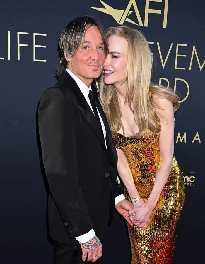 NIcole KIdman and Keith Urban pose together on a red carpet. One wears a suit, and the other wears a shimmering, form-fitting gown while whispering with a smile