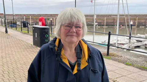 Liz Elmont is standing on a boardwalk by the harbour in Watchet. She is wearing a blue raincoat and is smiling at the camera.