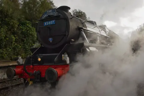 Paul Bradley The black steam locomotive "The Jacobite” stands on a railway track, surrounded by thick clouds of white steam. Its front features bright red buffers and metallic details, contrasting with the dark body of the train. Dense greenery lines the platform in the background.