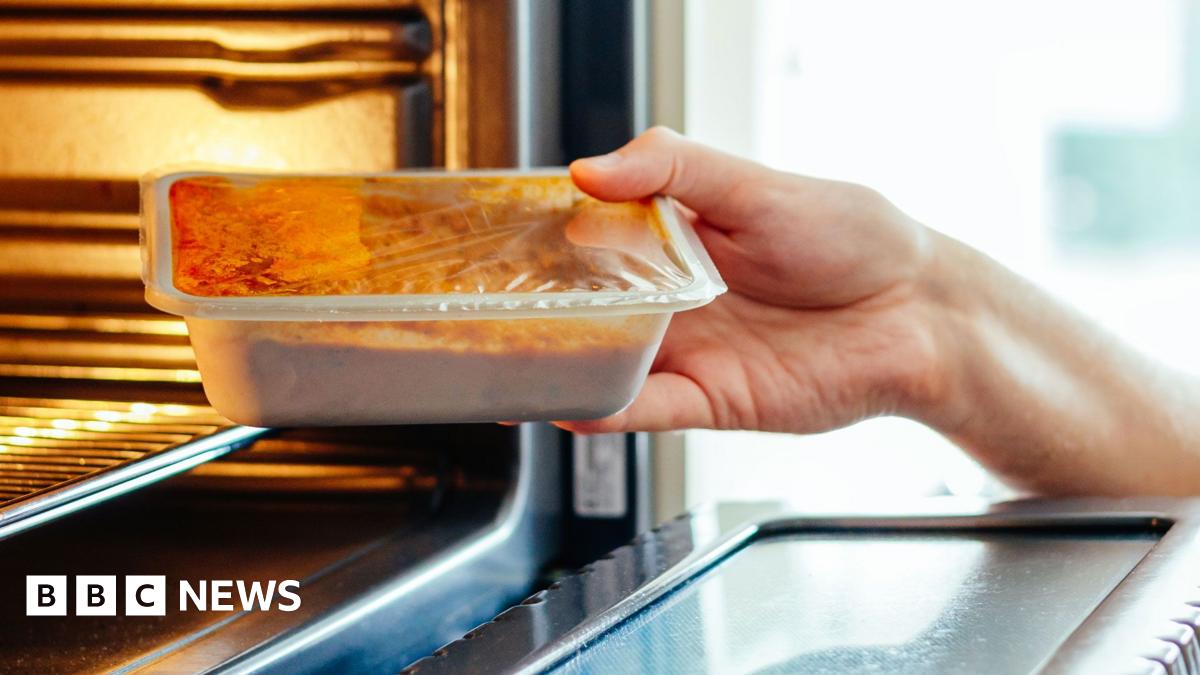 Looking down on a table full of plates of pizza, burgers in buns, onion rings, french fries and cola drinks, which are all examples of ultra-processed foods. Hands are reaching out and picking up the different foods.