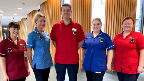 WHSCT A group of five health care staff wearing uniforms stand in a hospital corridor. A man is at the centre of the image, the other four are women.
