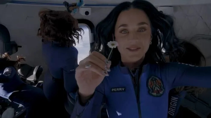 A person in a space capsule holds a white flower, wearing a blue flight suit labeled "Perry," with zero gravity evident by floating hair and objects
