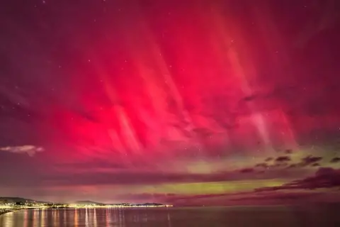 Ruth Davies | BBC Weather Watchers The lights from the coastline can be seen on the left with pink and green lights in the sky from the Northern Lights.