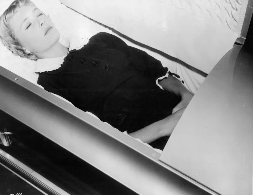 Christine White lying in an open casket, wearing a formal dress with a lace collar and buttons, with eyes closed and hands peacefully folded