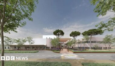 The picture shows a computer-generated design of the proposed new hospital or health campus at Overdale in Jersey. The building has a sleek, contemporary look with light-coloured walls and large windows.  The entrance is framed by landscaped greenery and tall trees, giving the site a campus-like atmosphere. A wide pedestrian pathway leads to the main entrance, and a road with a marked crossing runs in front of the building.