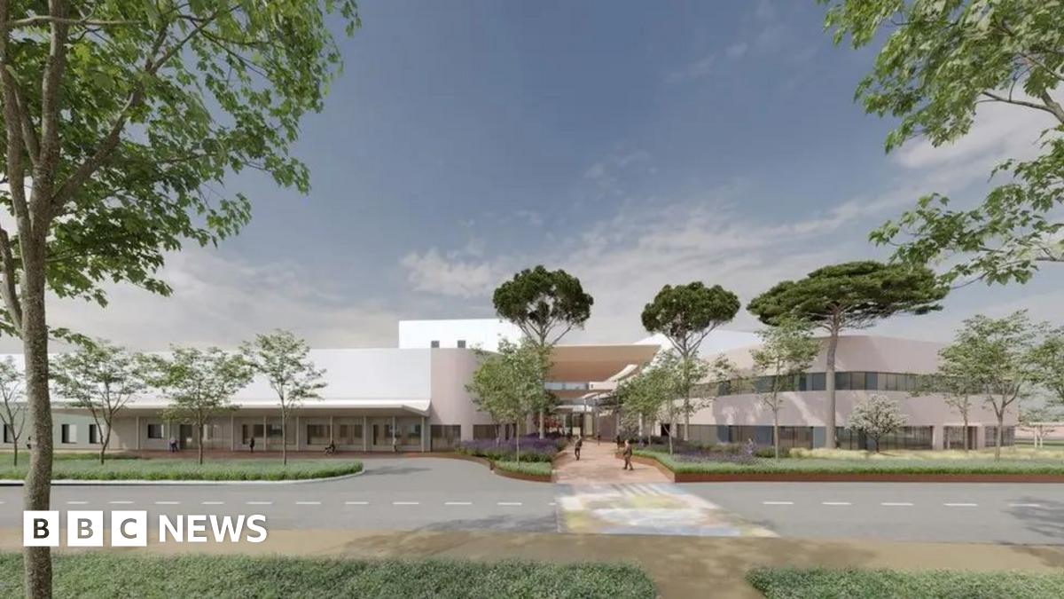 The picture shows a computer-generated design of the proposed new hospital or health campus at Overdale in Jersey. The building has a sleek, contemporary look with light-coloured walls and large windows.  The entrance is framed by landscaped greenery and tall trees, giving the site a campus-like atmosphere. A wide pedestrian pathway leads to the main entrance, and a road with a marked crossing runs in front of the building.