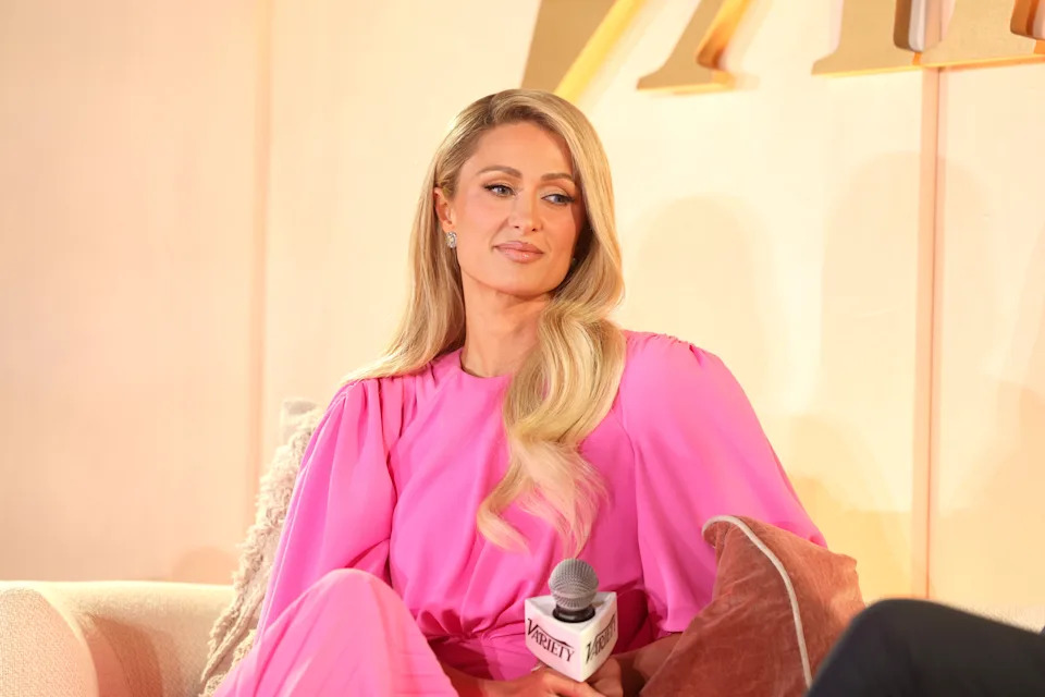 Paris Hilton in elegant dress seated, holding a microphone, appearing at an event or interview setup