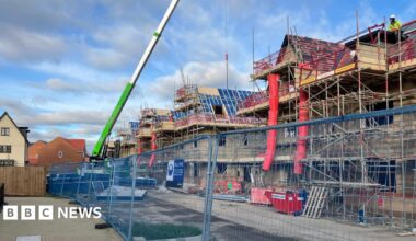 A metal fence with three-storey houses being built behind it, with scaffolding around it. There is a crane, blue skies and white clouds above.