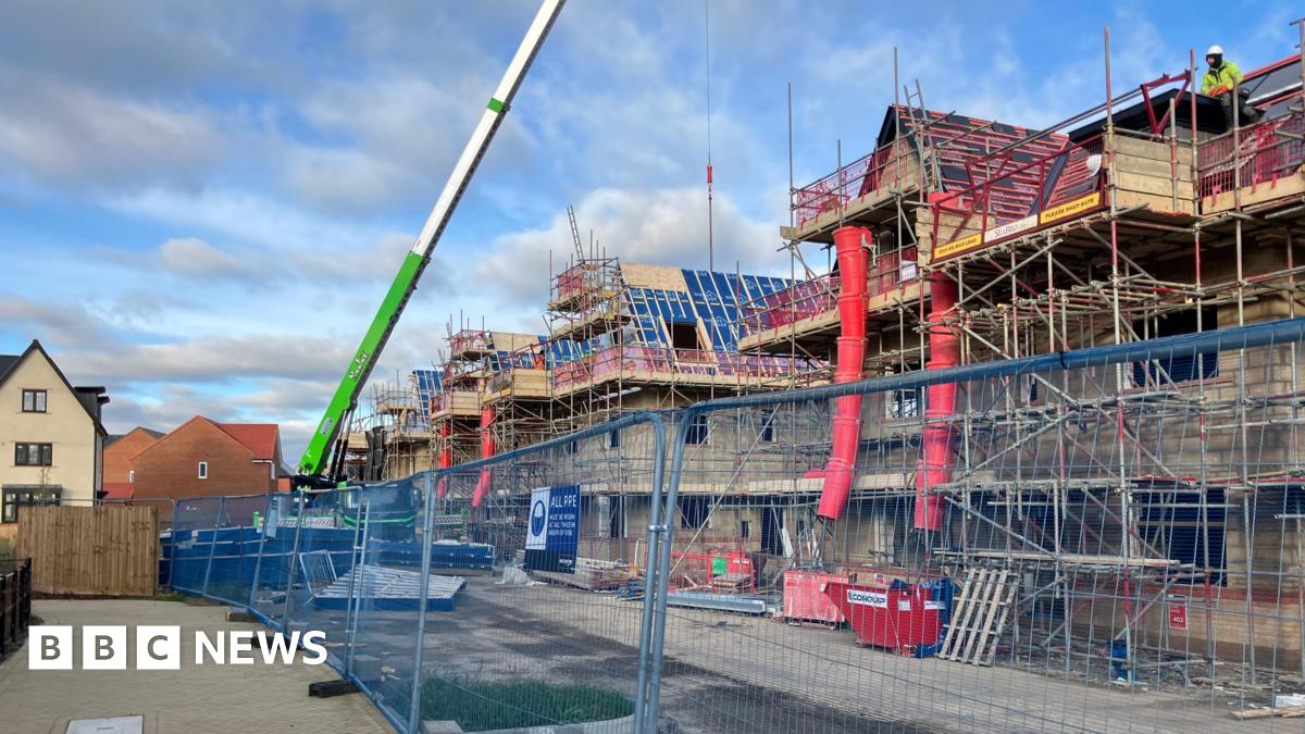 A metal fence with three-storey houses being built behind it, with scaffolding around it. There is a crane, blue skies and white clouds above.