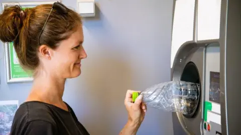 Getty Images Image of a woman with dark hair piled up in a bun, with dark sunglasses on top of her head, posting a plastic water bottle into a large machine roughly the shape of a traditional drinks vending machine