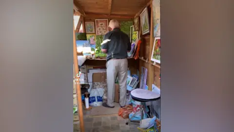 Jo Menzies/TikTok A grab of a TikTok video showing Mr Rose with his back to the camera, standing painting in his shed which is adorned with other paintings.
