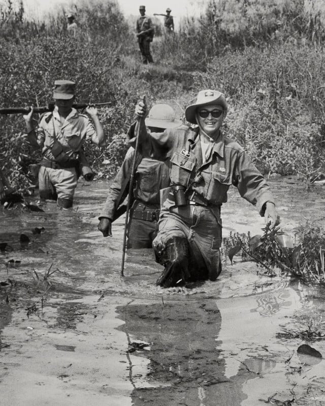 Three soldiers wade through swampy water in a grassy area, wearing military uniforms and gear. One in front smiles at the camera, while two others follow closely behind. More soldiers are visible in the background.