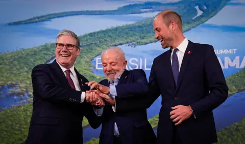 PA Media Sir Keir Starmer (l), Brazilian President Lula (c) and the Prince of Wales (r) clasp hands in a triple handshake, laughing together, on the podium at COP30, in Belem. The trio wear dark suits, with Sir Keir and Prince Wiliam wearing shirt and tie, and Lula with an open-neck shirt. 