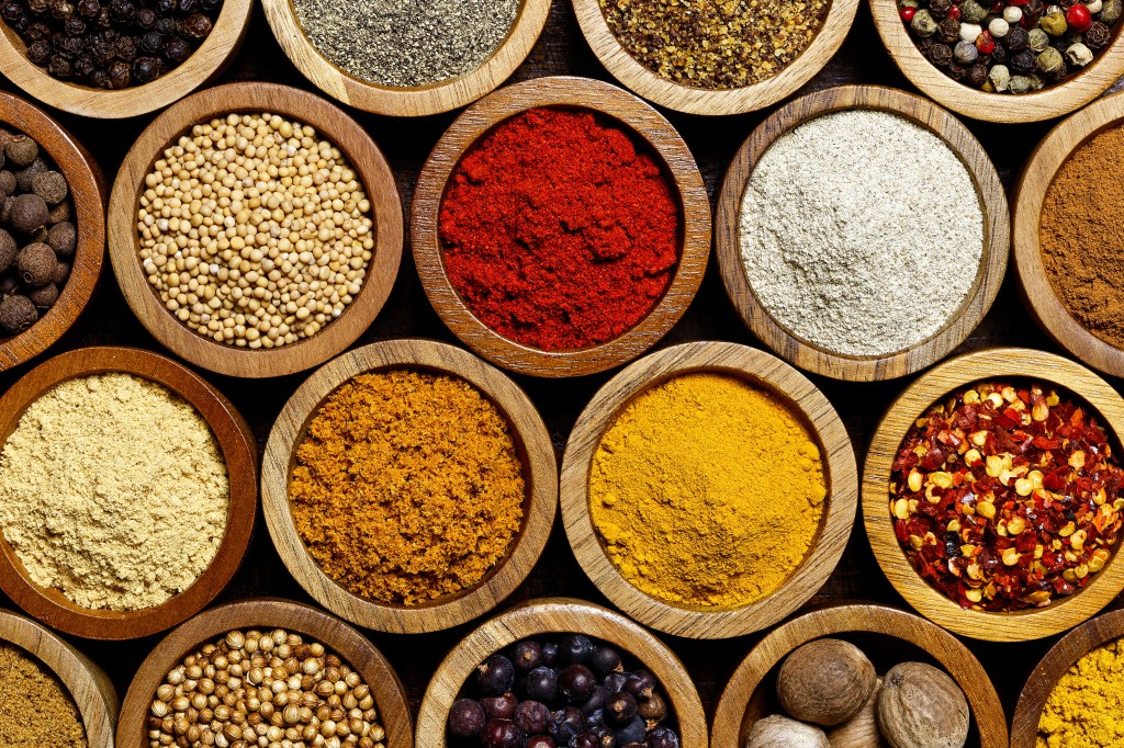 Various spices arranged in small wooden bowls, including whole black peppercorns, mustard seeds, paprika, white pepper, allspice, ginger powder, curry powder, turmeric, red pepper flakes, ground cumin, coriander seeds, juniper berries, and whole nutmeg.