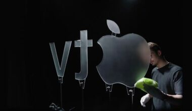A person wearing gloves cleans a large glass Apple logo next to "vt" symbols, all standing upright against a dark background.