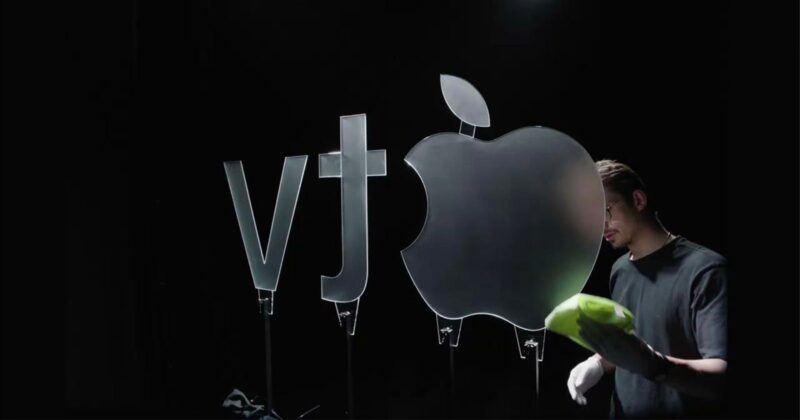 A person wearing gloves cleans a large glass Apple logo next to "vt" symbols, all standing upright against a dark background.